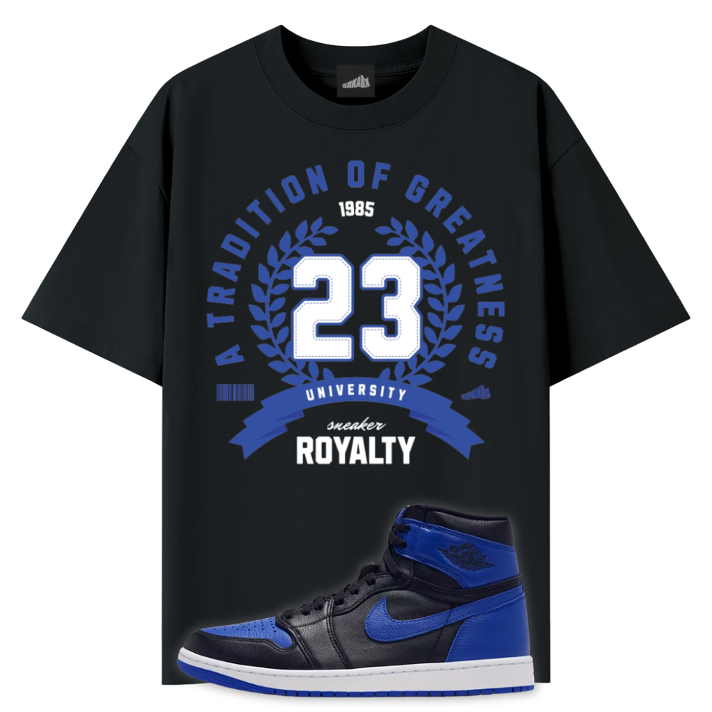A black graphic T-shirt featuring a blue and white '23' laurel wreath design with the text 'A Tradition of Greatness', '1985', 'University', 'Sneaker', and 'Royalty'. A blue, black, and white Air Jordan 1 Royal high-top sneaker is shown overlapping the lower right portion of the shirt on a plain white background.