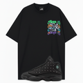A black oversized short-sleeve t-shirt with a colorful graphic on the left chest and a black Air Jordan 13 Wings sneaker overlaid at the bottom, shown on a light gray background.