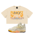 A cream-colored cropped t-shirt featuring a graphic print with the words 'Paradise of' in orange and blue, accompanied by bird illustrations, shown alongside a matching cream, orange, and yellow Air Jordan 4 Bird of Paradise sneaker on a plain white background.