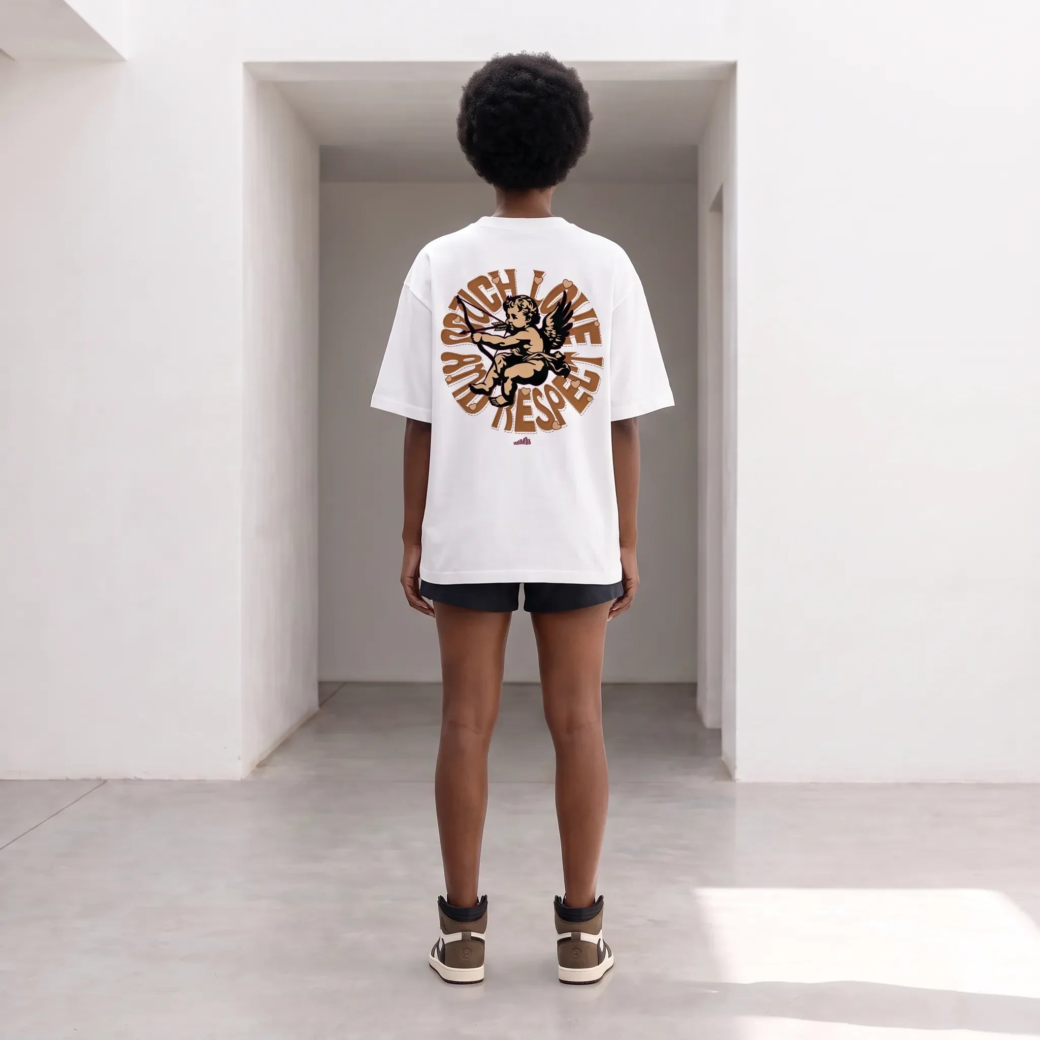 A model viewed from behind wearing an oversized white t-shirt with a large brown graphic print on the back, paired with black shorts and brown sneakers, standing in a minimalist white hallway.