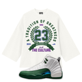 An off-white short-sleeve crop top featuring a green and purple graphic print on the front with the number 23, laurel wreath, and the text 'A TRADITION OF GREATNESS' and 'built THE CULTURE' with a Air Jordan 12 Bucks sneaker in the front.
