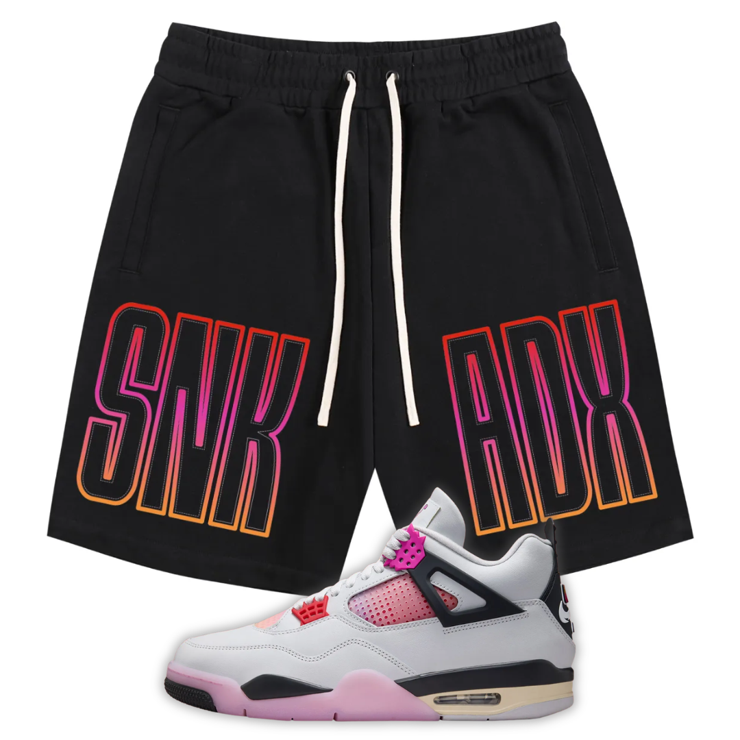 Black athletic shorts with white drawstrings and large 'SNKADX' text printed on the sides in a pink-to-orange gradient, shown with a white, pink, and black Air Jordan 4 Comic sneaker at the bottom.