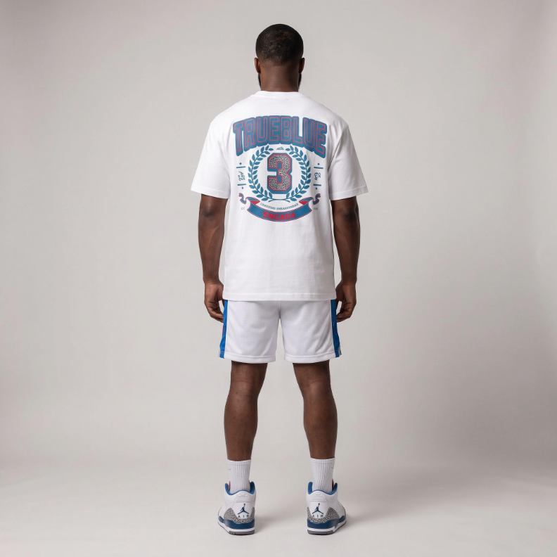 A man viewed from the back wearing a white t-shirt with a large blue and red graphic on the back, white athletic shorts with blue side stripes, white socks, and Jordan 3 True Blue sneakers, set against a plain light gray studio background.