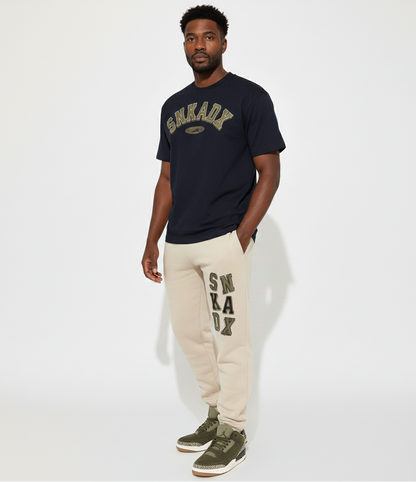 Man wearing black t-shirt and beige sweatpants with 'SNKADX' branding on a white background